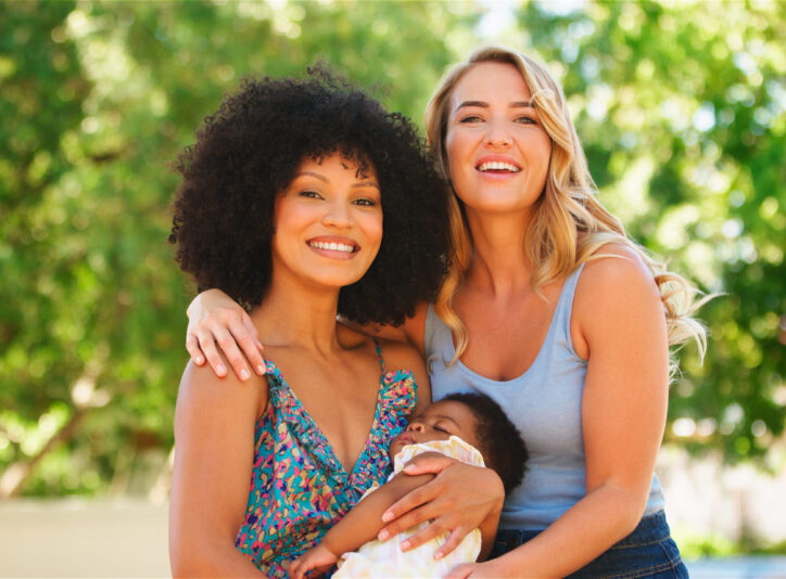 Two women happily pose together with a baby in a lush green setting.